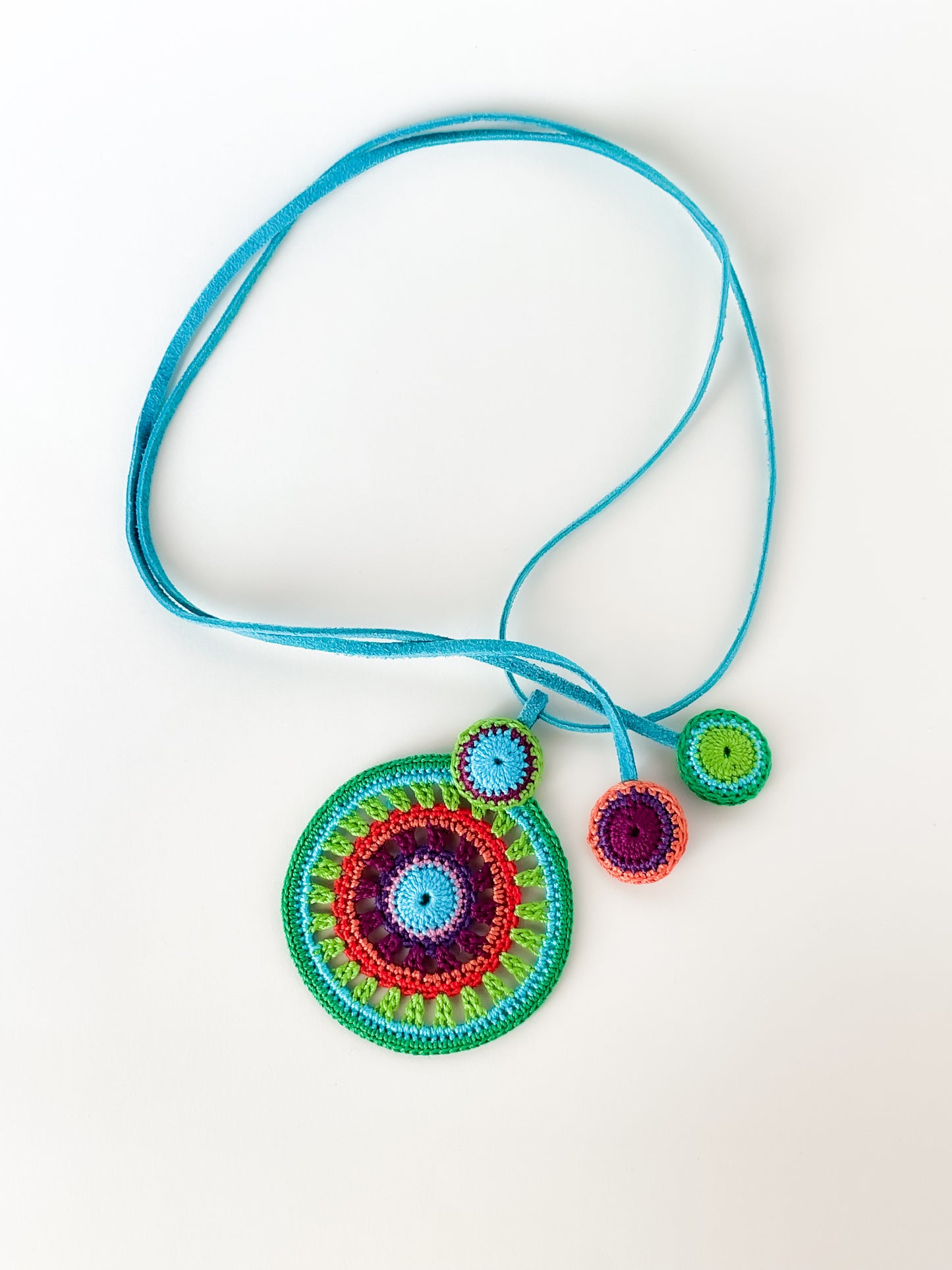Hand-crocheted necklace featuring bold concentric circles in turquoise, red, purple and green tones, with a matching blue suede cord. Unique artisan jewelry piece.