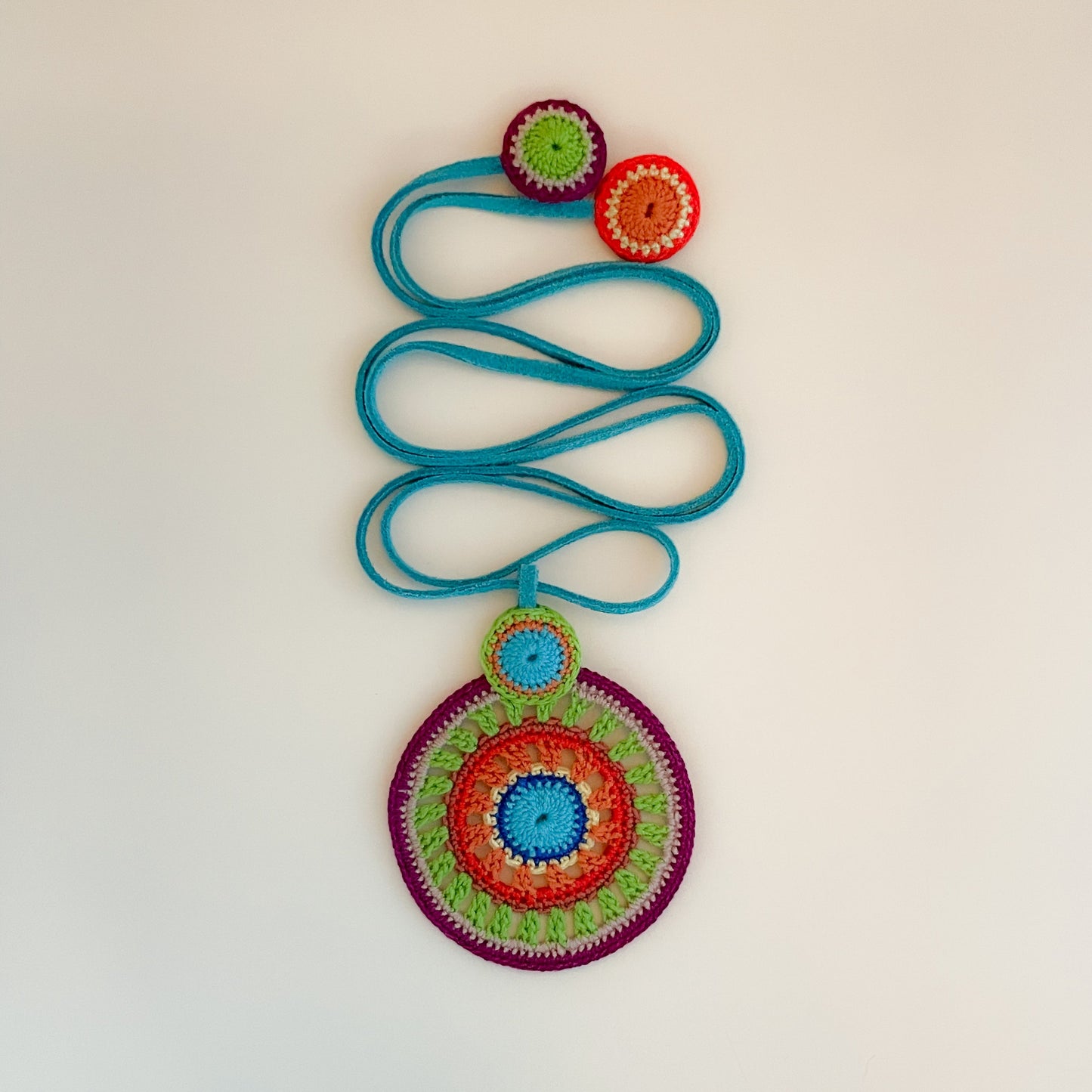 Artisan crochet necklace in bold colors of blue, green, orange and deep purple, featuring circular motifs on a blue suede cord. One-of-a-kind wearable art.