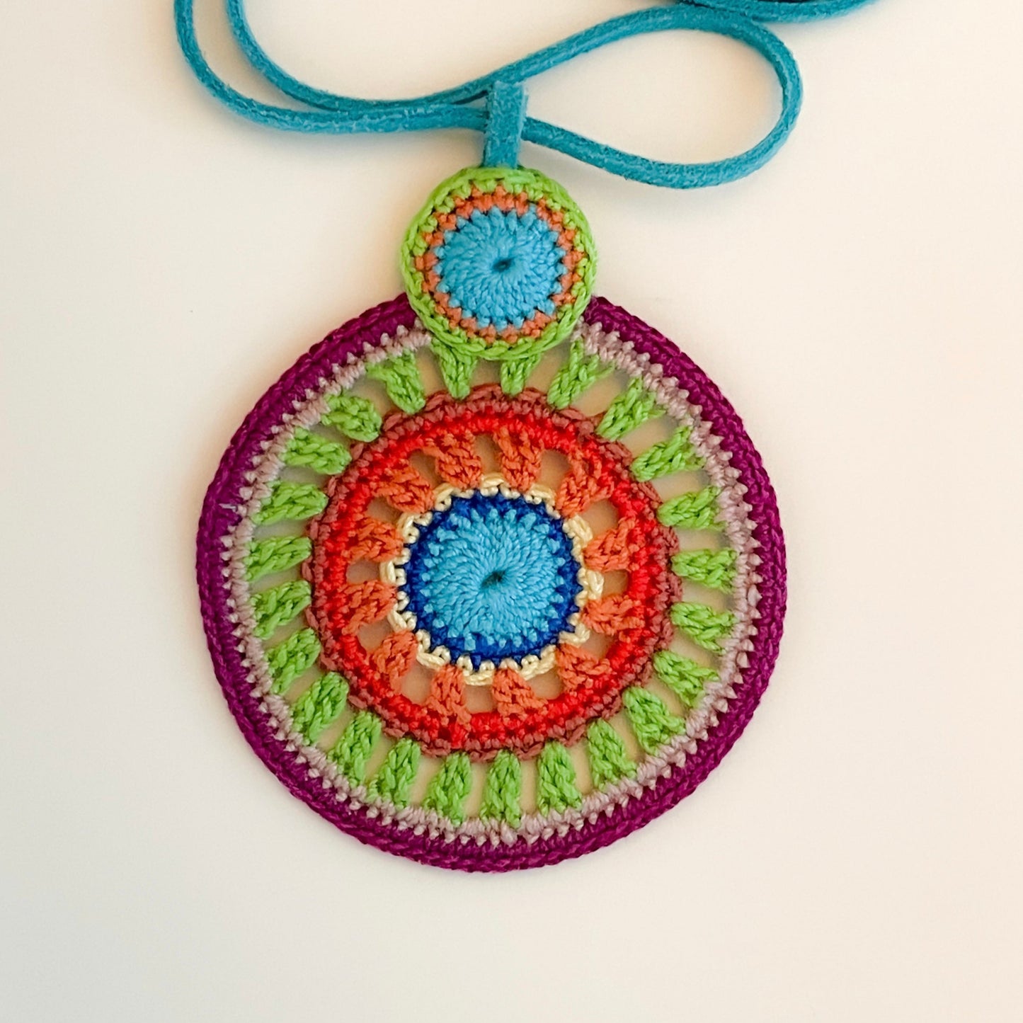 Close-up of intricate crochet work on colorful pendant necklace, highlighting detailed handmade craftsmanship and vibrant thread combinations in purple, green, red and blue tones.