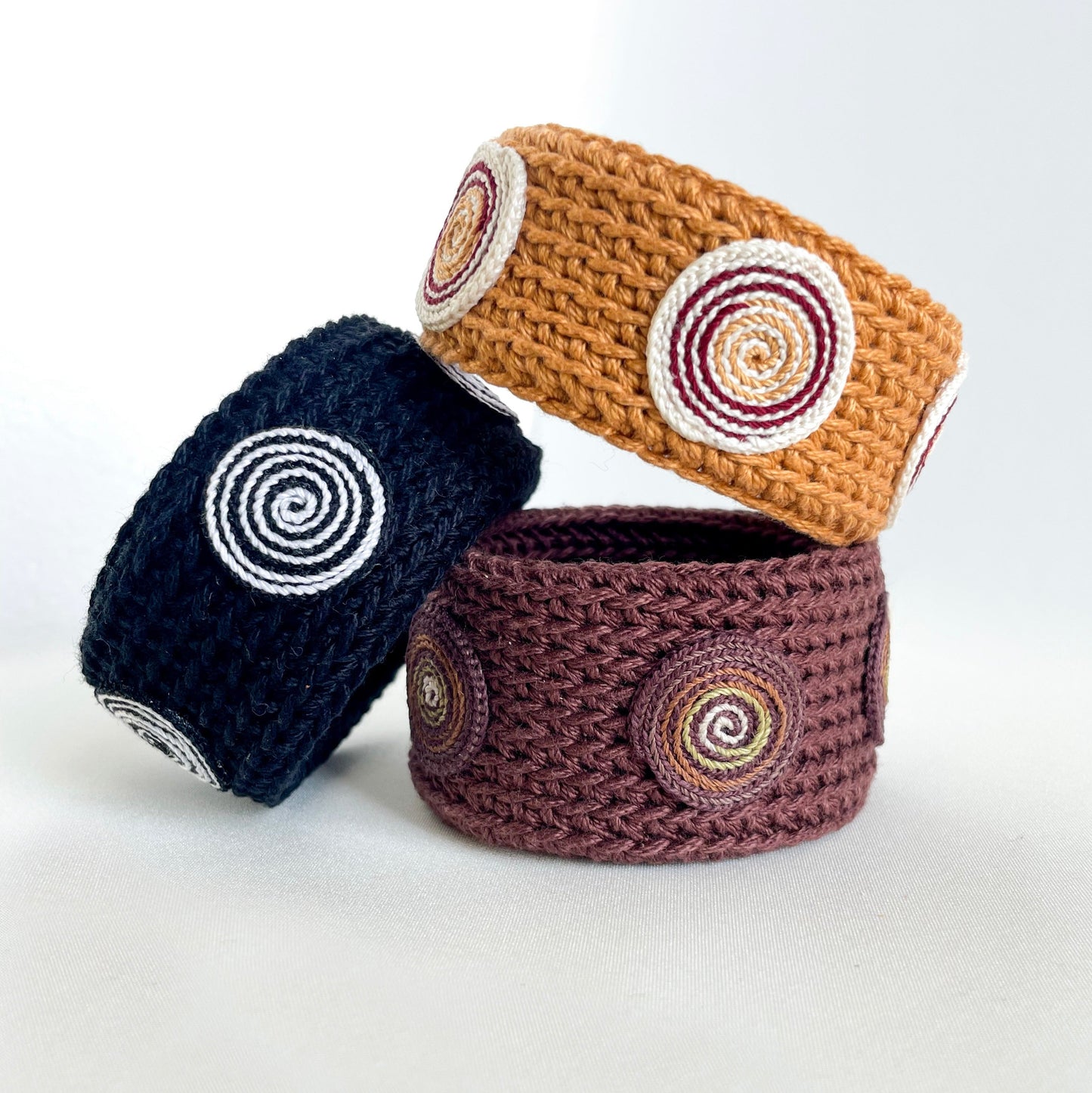 Three crocheted baskets in navy, brown, and beige with circular patterns on a white background. Set of three Spiralis crochet bracelets in black, brown, and amber tones with spiral designs.