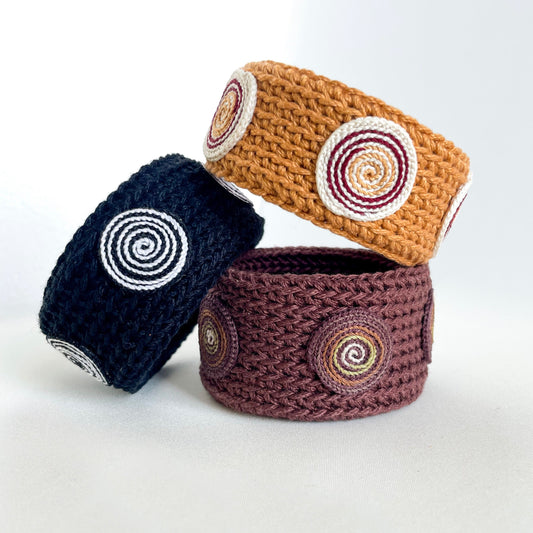 Three crocheted baskets in navy, brown, and beige with circular patterns on a white background. Set of three Spiralis crochet bracelets in black, brown, and amber tones with spiral designs.