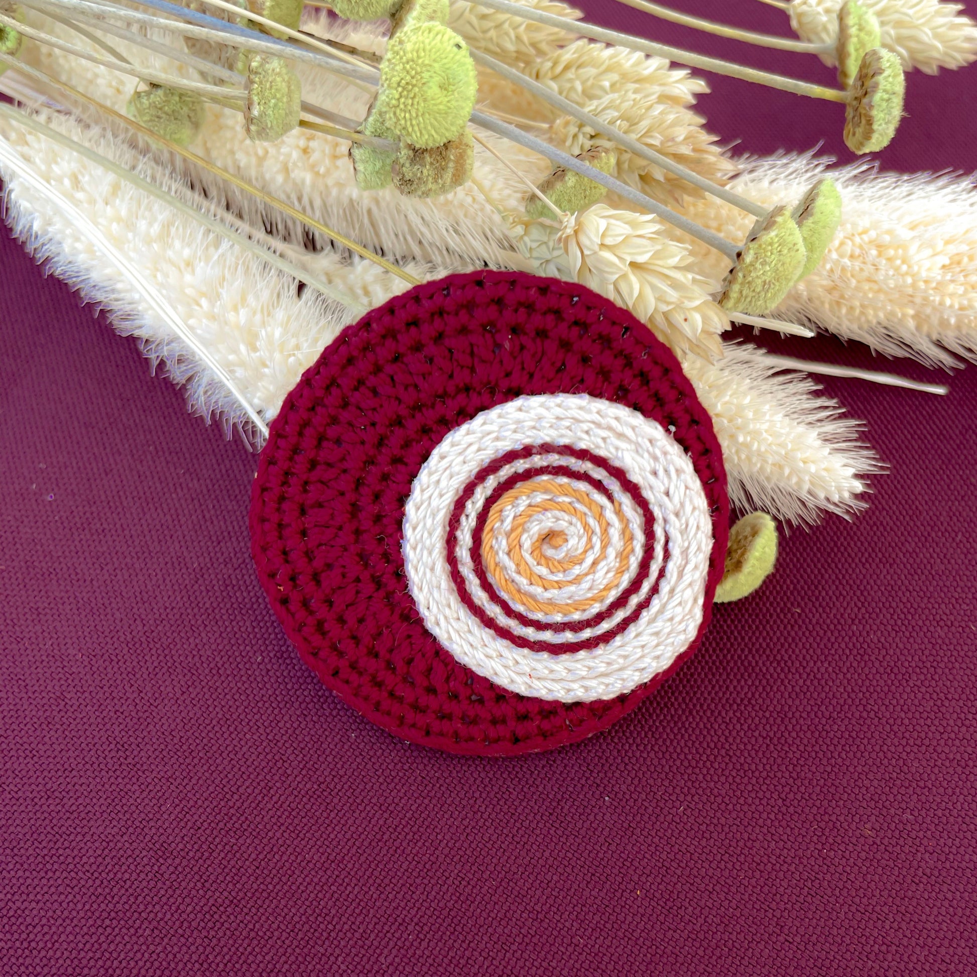 Crocheted circular Pin with concentric circles on a burgundy surface with dried plants.