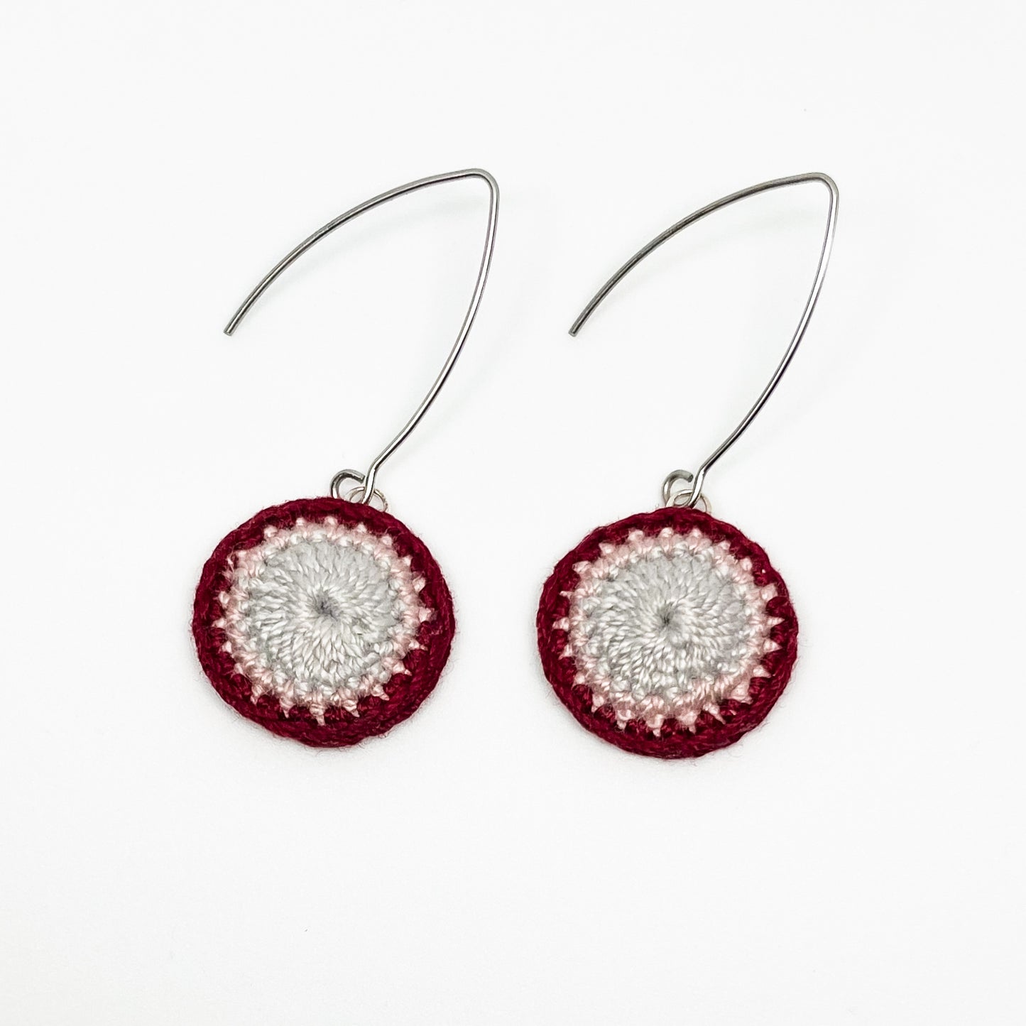 A close-up of small round crochet earrings in currant and cloud colors.