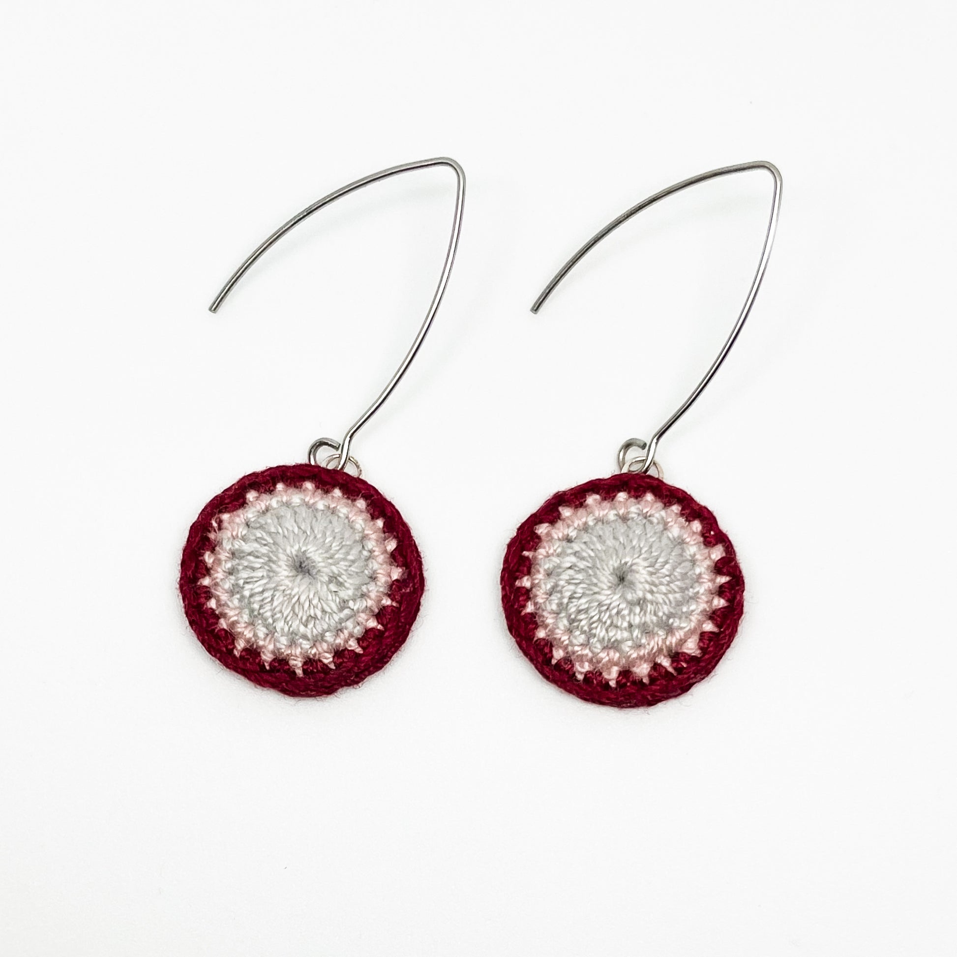 A close-up of small round crochet earrings in currant and cloud colors.