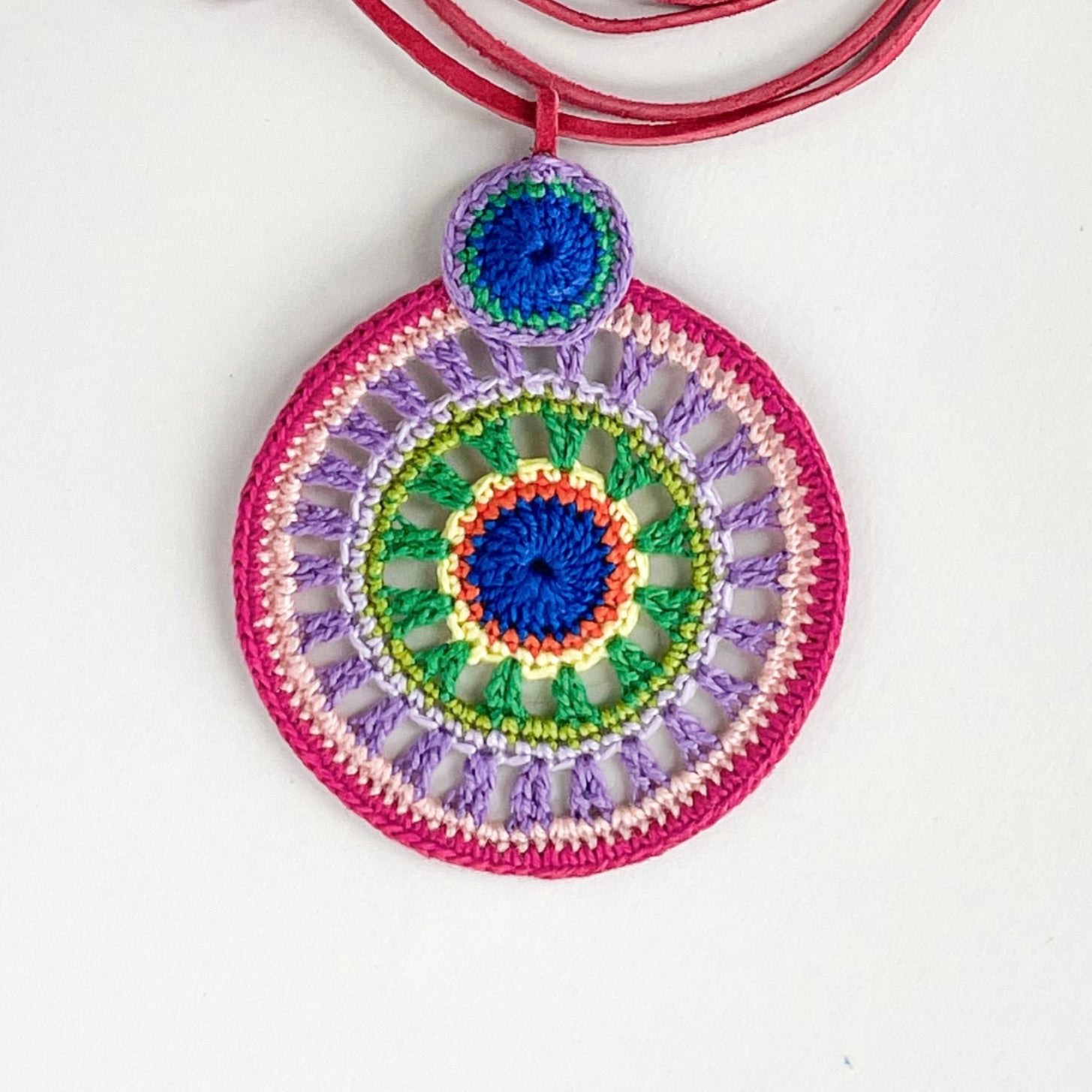 Close-up of intricate crochet work on colorful pendant necklace, highlighting detailed handmade craftsmanship and vibrant thread combinations iof lilac, pink, blue and green.