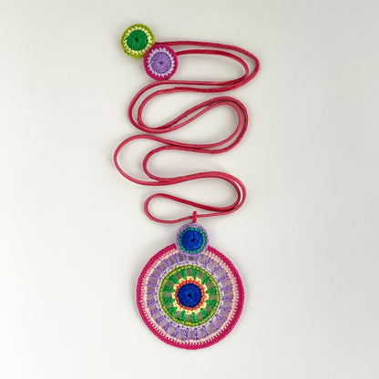 Feminine crochet pendant necklace in pink, purple, green and blue shades with deep pink cord – handcrafted statement jewelry on white background.
