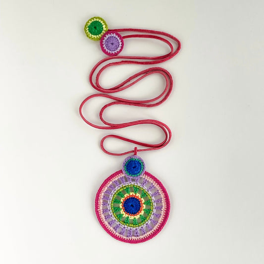 Feminine crochet pendant necklace in pink, purple, green and blue shades with deep pink cord – handcrafted statement jewelry on white background.