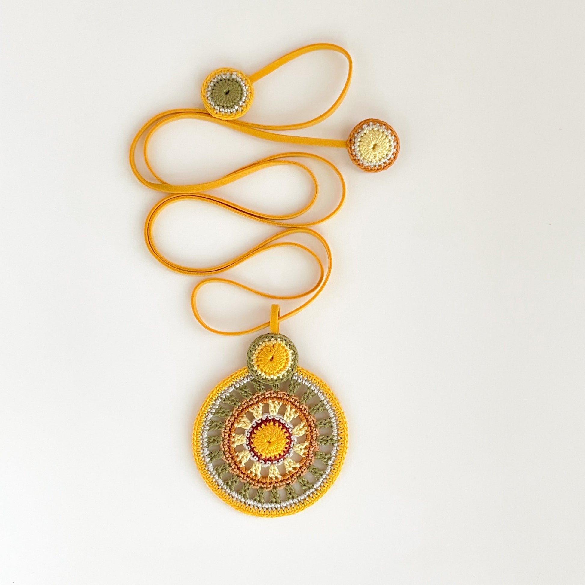 Handmade colorful crochet necklace with concentric circles in warm tropical tones of orange, yellow, olive green and cream, finished with a mustard suede cord– front view on white background.