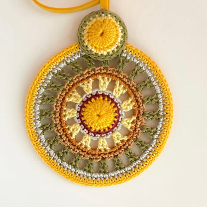 Close-up of intricate crochet work on colorful pendant necklace, highlighting detailed handmade craftsmanship and vibrant thread combinations in warm tropical tones of orange, yellow, olive green and cream.