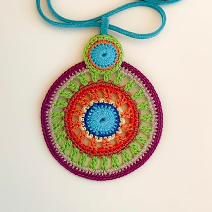 Close-up of intricate crochet work on colorful pendant necklace, highlighting detailed handmade craftsmanship and vibrant thread combinations in purple, green, red and blue tones.