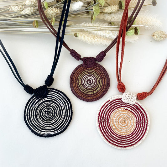 Three crocheted necklaces with spiral design on a white surface with dried plants.

