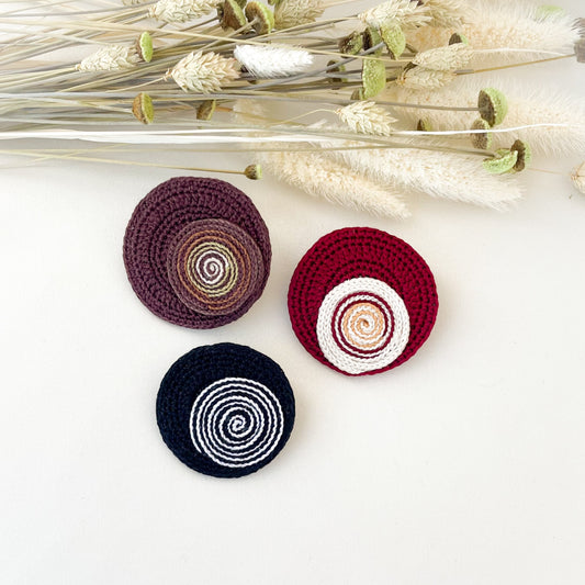 Three crocheted circular pins in different colors on a white surface with dried flowers.