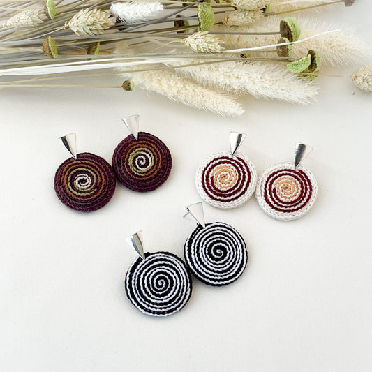 Three sets of circular earrings with spiral patterns on a white background with dried plants.
