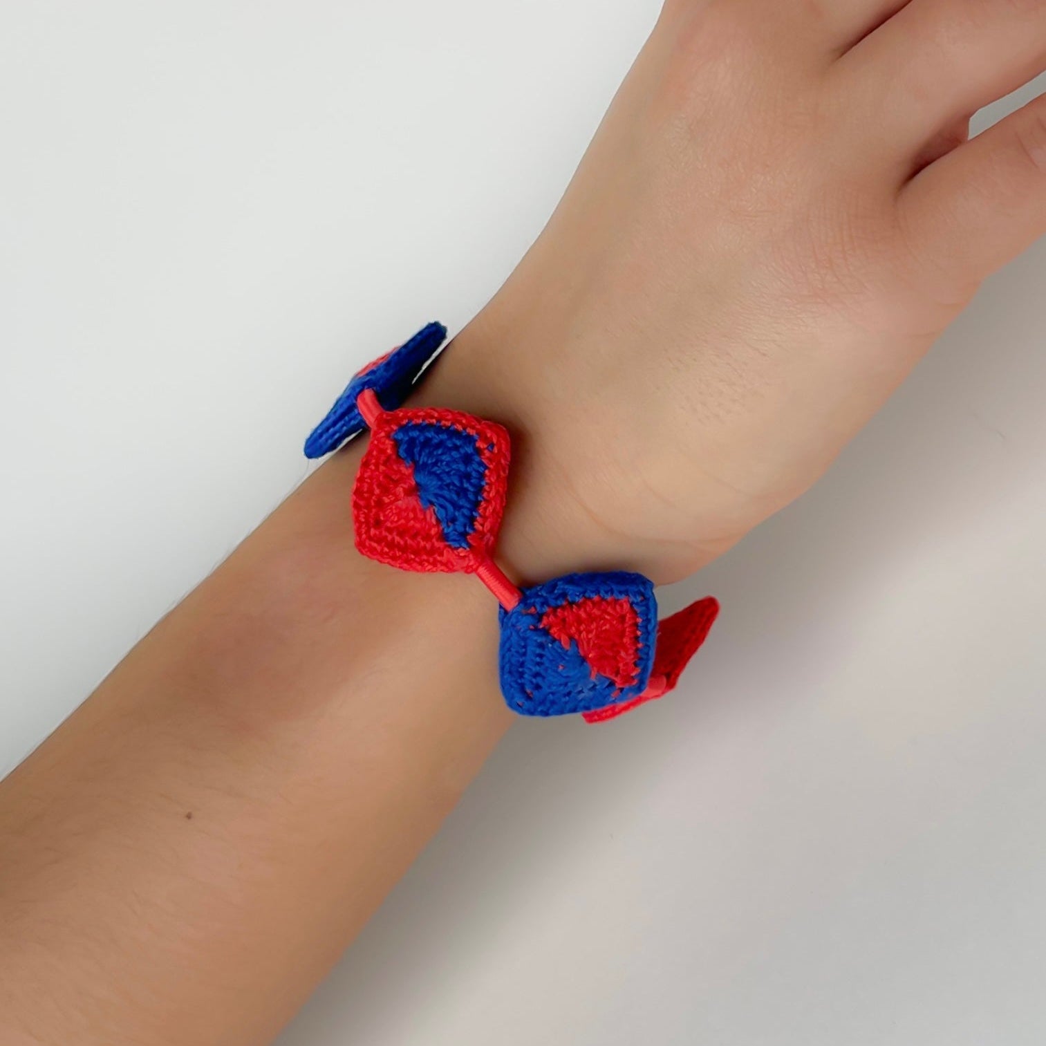 Hand wearing a Tessera crochet bracelet by Crochet by Heart showing the handmade geometric crochet tiles in red and blue on a plain background
