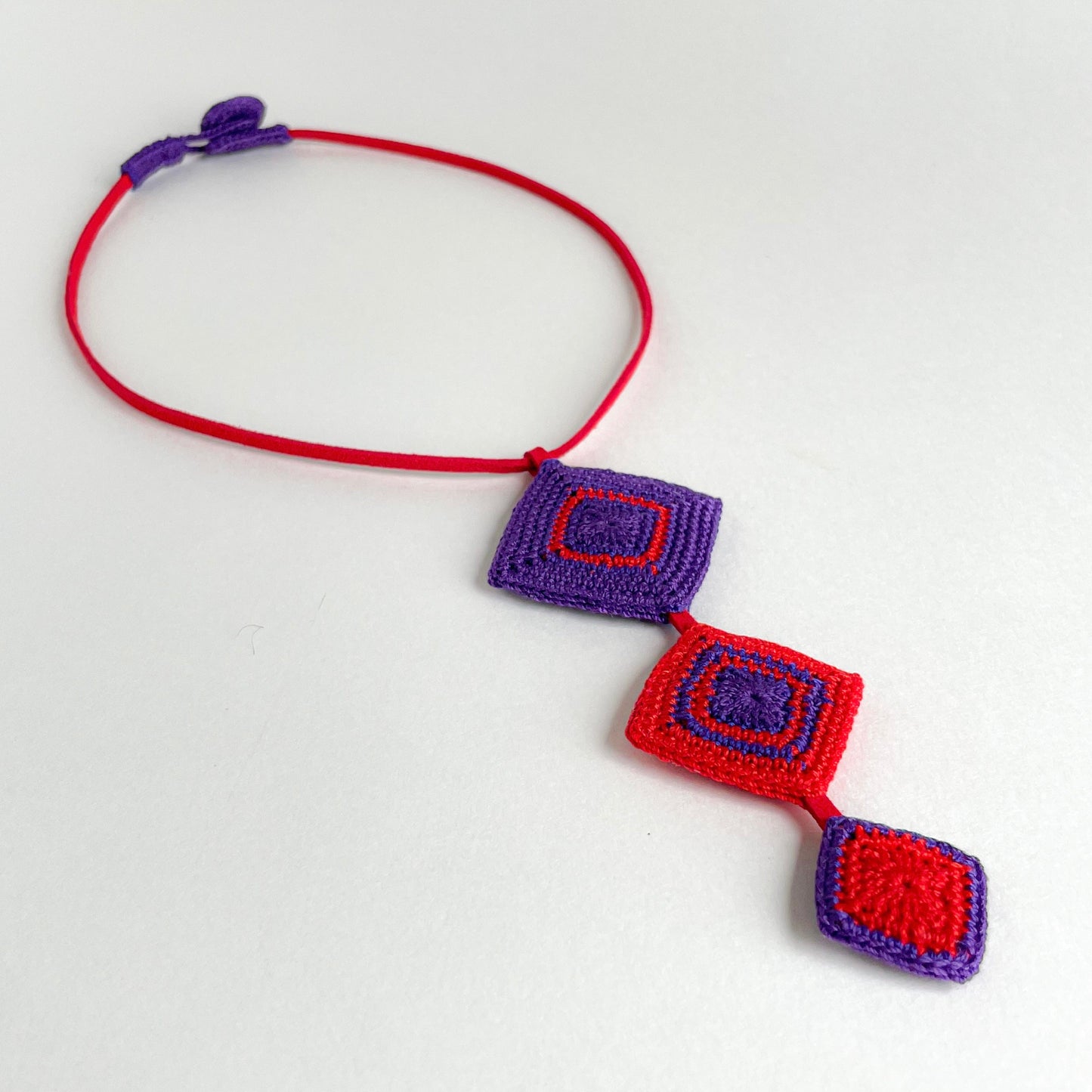 Front view of the Tessera geometric crochet necklace by Crochet by Heart featuring red and purple diamond motifs on a red cord.