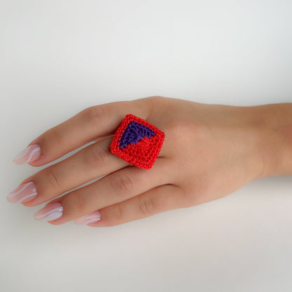 Hand wearing a red and violet Tessera crochet ring on a white background.