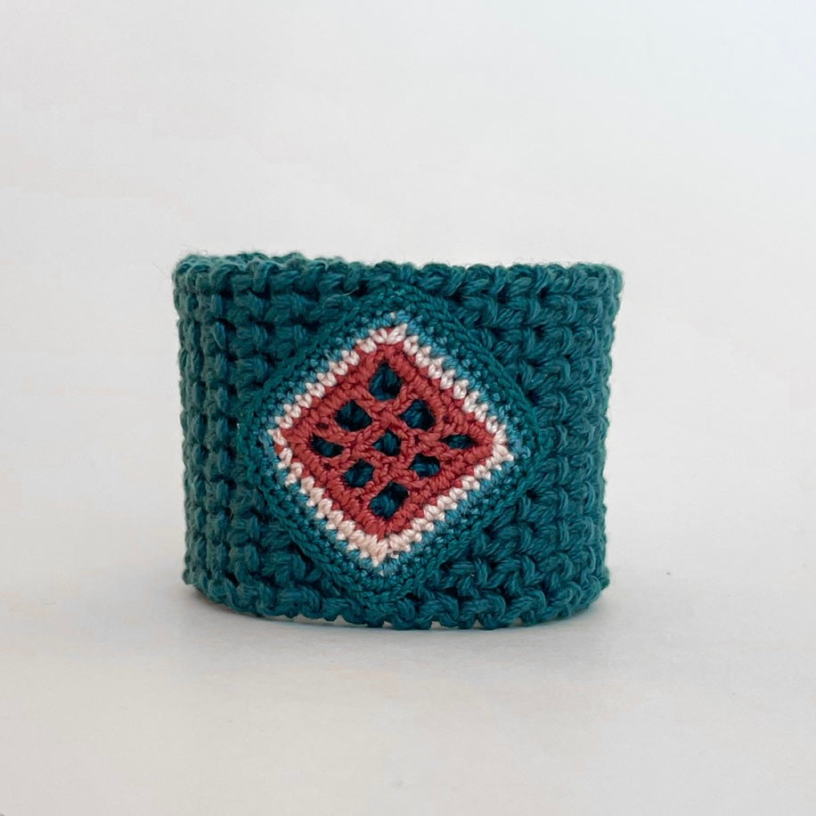 Front view of the TRAMA teal crochet cuff bracelet with a clay openwork motif bordered in ivory,  blue and teal.