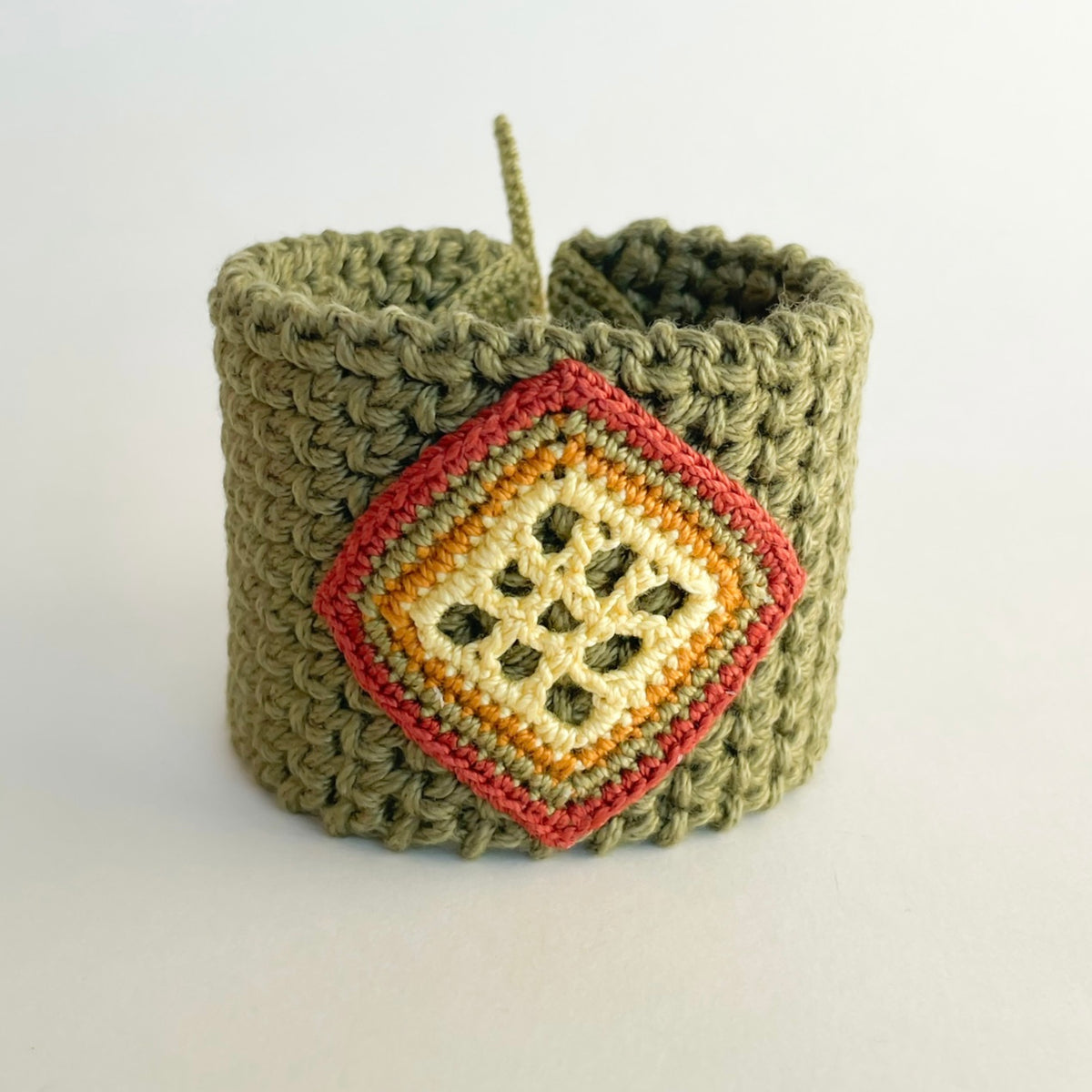 A handmade olive green crocheted cuff bracelet with a diamond-shaped front charm featuring yellow butter, ginger, moss green and cooper stripes.