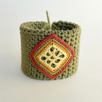 A handmade olive green crocheted cuff bracelet with a diamond-shaped front charm featuring yellow butter, ginger, moss green and cooper stripes.