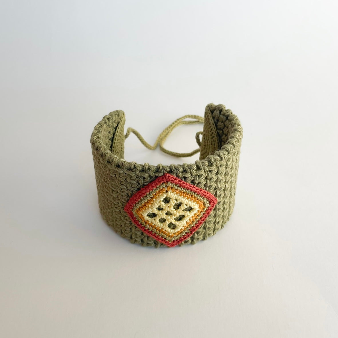 A handmade olive green crocheted cuff bracelet with a decorative diamond-shaped patch featuring yellow butter, ginger, moss green and cooper colors, showing the tie cords behind, set against a plain light gray background.