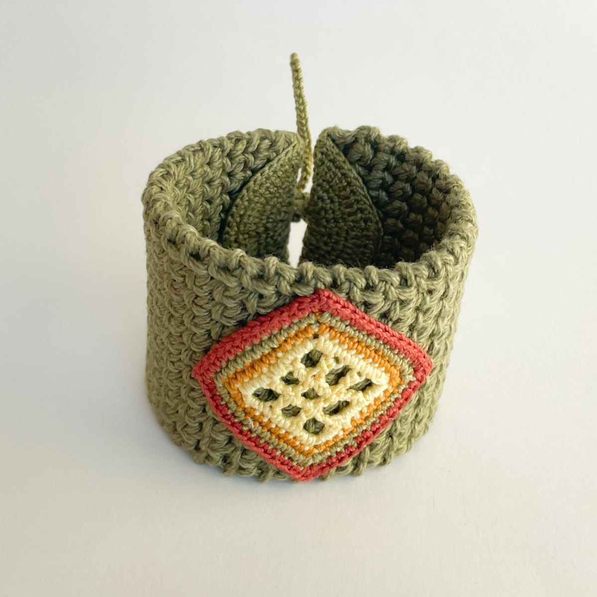 Angled view of the TRAMA moss green crochet cuff bracelet, showing its textured stitches, colourful diamond motif and tie closure.
