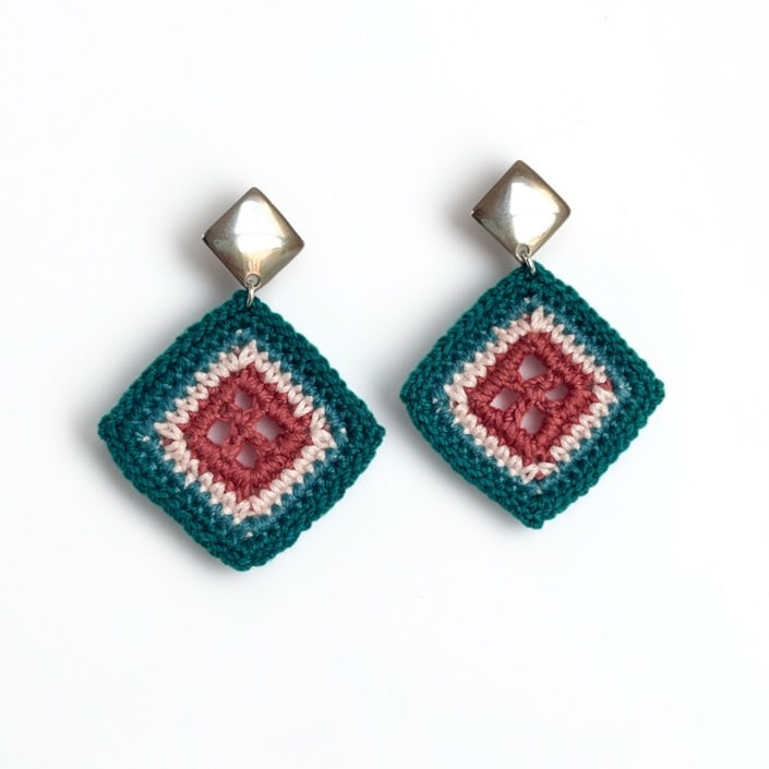 A pair of handmade crochet dangle earrings with a geometric diamond design. The earrings feature a dark teal outer crochet border, a clay center crochet square, and a cream inner crochet square, hanging from shiny silver-toned metal studs.