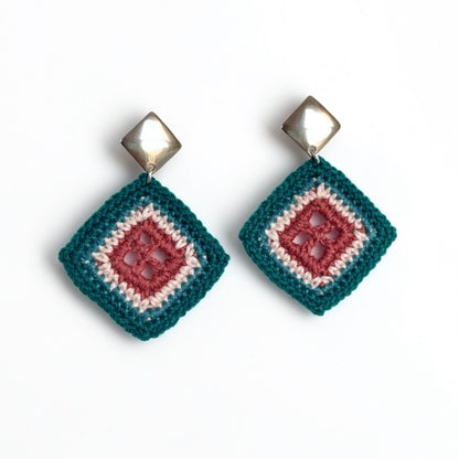 A pair of handmade crochet dangle earrings with a geometric diamond design. The earrings feature a dark teal outer crochet border, a clay center crochet square, and a cream inner crochet square, hanging from shiny silver-toned metal studs.
