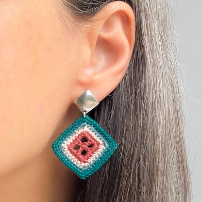 A close-up of a person's ear wearing a handmade crocheted dangle earring. The earring features a silver-toned metal top and a square drop with concentric bands of teal, dusty rose, and clay yarn.
