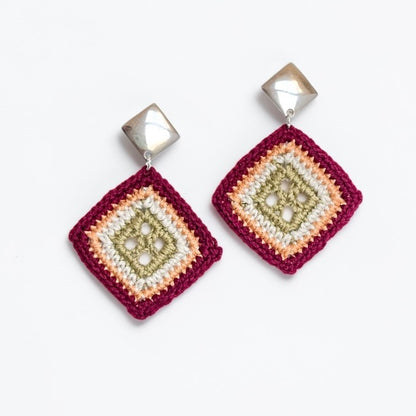 A pair of handmade square dangle earrings featuring a multi-colored crocheted pattern in burgundy, peach, cream, and olive green, suspended from faceted silver-toned cube studs.