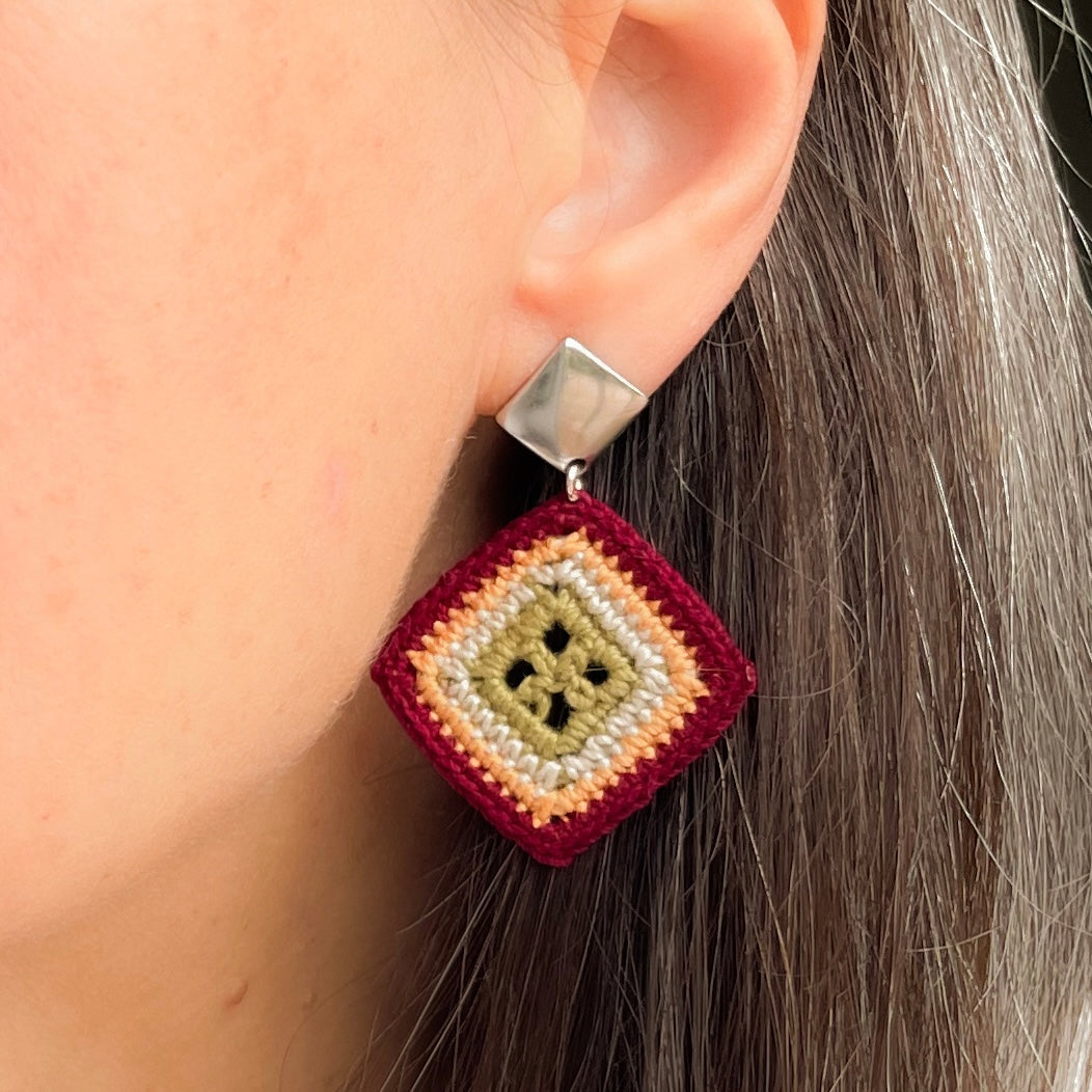 A close-up of a person's ear wearing a handmade geometric dangle earring. The earring features a silver-toned square stud and a diamond-shaped drop made of layered, knotted yarn in burgundy, marigold, cream and olive green.