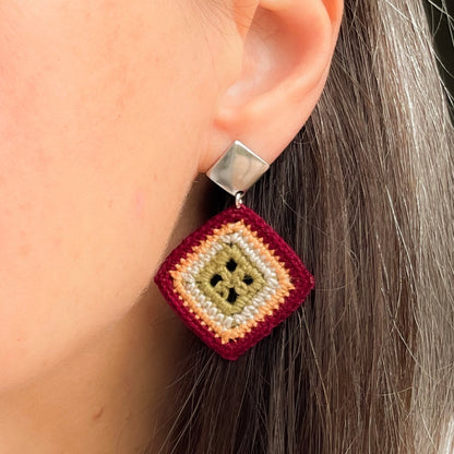 A close-up of a person's ear wearing a handmade geometric dangle earring. The earring features a silver-toned square stud and a diamond-shaped drop made of layered, knotted yarn in burgundy, marigold, cream and olive green.