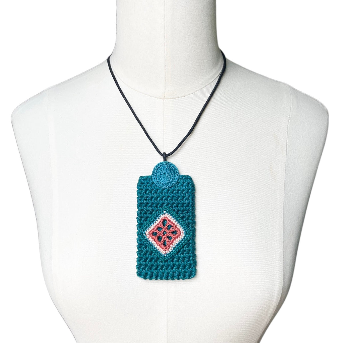 A teal crocheted necklace pendent with a clay and blue jade diamond pattern, hanging on a black cord from a white mannequin torso.