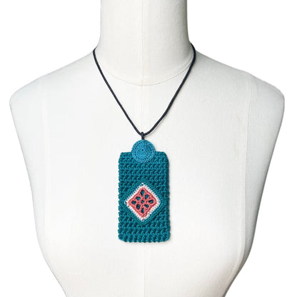 A teal crocheted necklace pendent with a clay and blue jade diamond pattern, hanging on a black cord from a white mannequin torso.