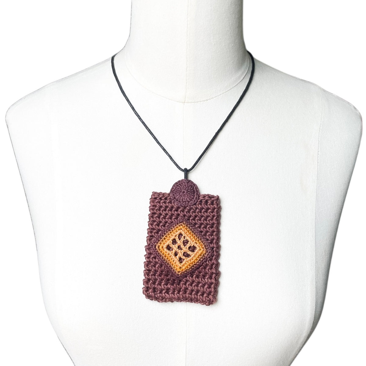 TRAMA brown rectangular crochet pendant with a marigold, ginger and chocolate diamond motif, hanging from a black cord on a white mannequin.