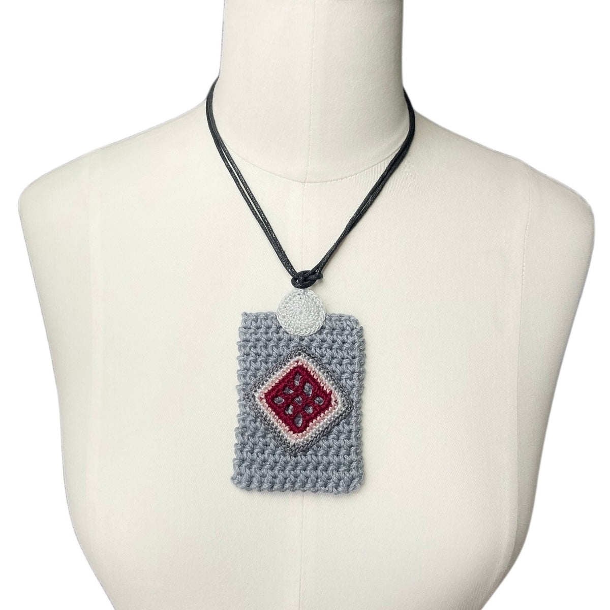 A crocheted gray rectangular holder necklace with a burgundy diamond patterned center, displayed on a cream-colored mannequin torso against a white background.
