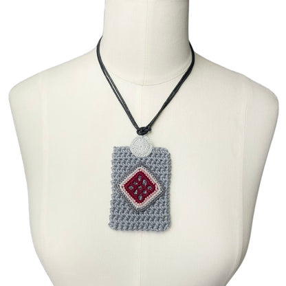 A crocheted gray rectangular holder necklace with a burgundy diamond patterned center, displayed on a cream-colored mannequin torso against a white background.