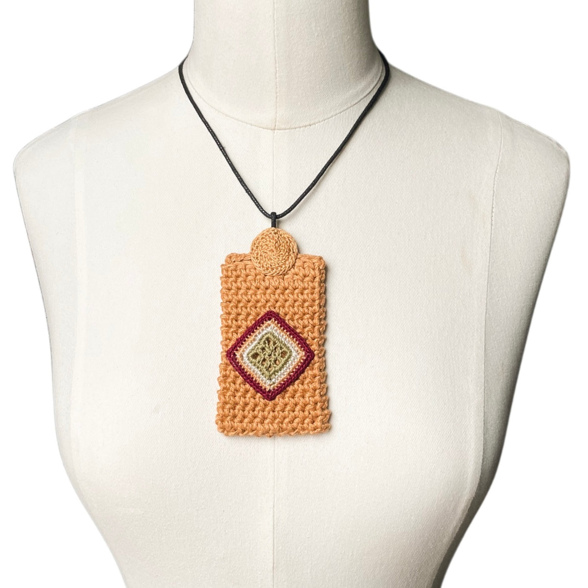 A crocheted marigold pendant necklace with a geometric diamond pattern in burgundy, beige, and green, displayed on a cream-colored mannequin torso.