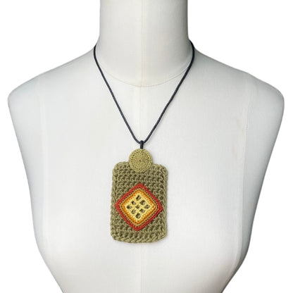 A crocheted rectangular pendant necklace displayed on a white mannequin torso. The pendant is olive green with a copper and yellow diamond pattern in the center hanging from a black cord on a white mannequin.