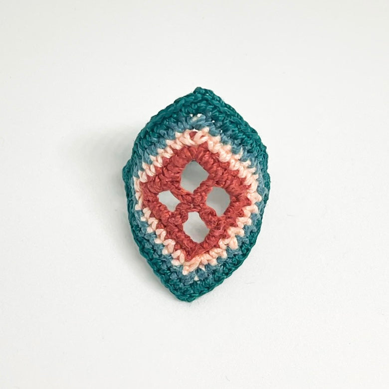 TRAMA teal crochet ring with an elongated diamond shape, clay openwork centre and pale cream border on a white background.