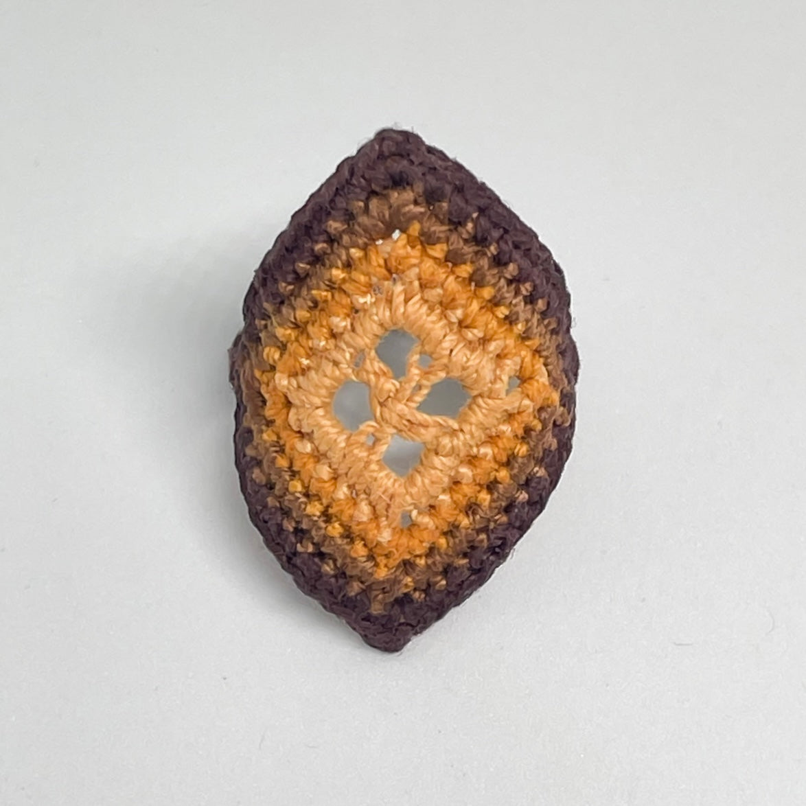 TRAMA chocolate crochet ring with an elongated diamond shape, marigold openwork centre and coconut border on a white background.