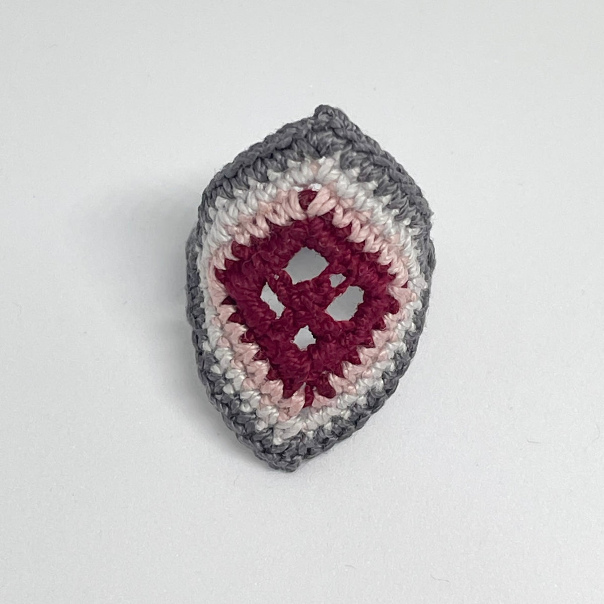 TRAMA gray crochet ring with an elongated diamond shape, burgundy openwork centre and rose border on a white background.