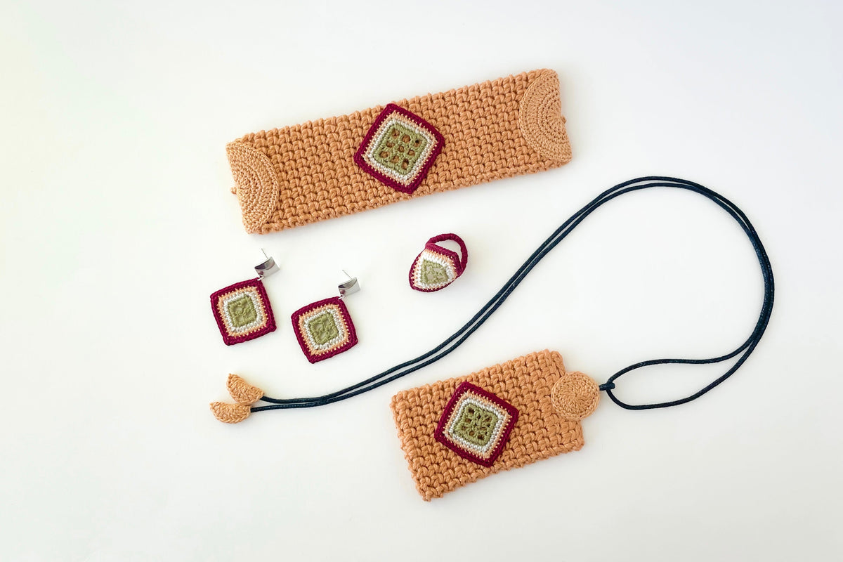 A coordinated handmade crochet jewelry and accessory set laid out on a white background, including a long rectangular pendant with a black cord, a matching bracelet,  a headband, two square earrings, and a small ring. The crochet fabric is marigold with burgundy and moss green square details.